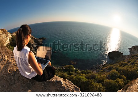 Young business woman working at the computer on the beach on the rock face. Young girl downshifter working at a laptop at sunset or sunrise on the top of the mountain to the sea, working day.