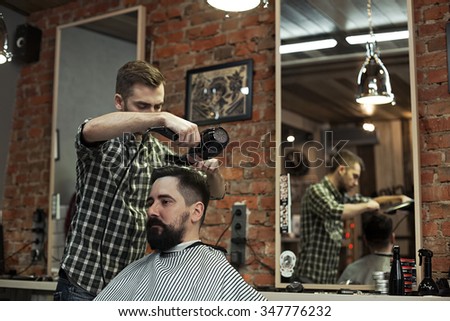 Image of cutting hair in a man\'s barber shop. The working day in Barbershop, stylish hipsters client with beard and mustache Stylist makes hair styling hair dryer.