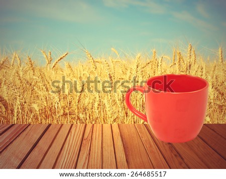 Red tea mug on wooden surface against of golden wheat ears and blue sky.
