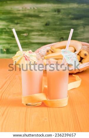 banana juice in two cups with straws and umbrellas on a yellow table with bananas on background