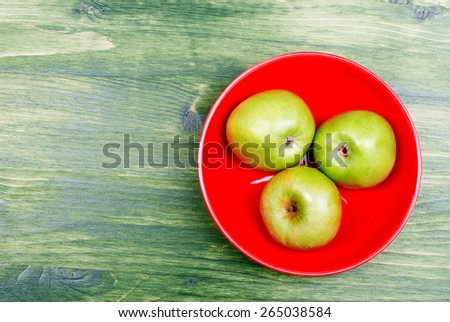 Three green apple in the red plate on the right edge of the green table, top view