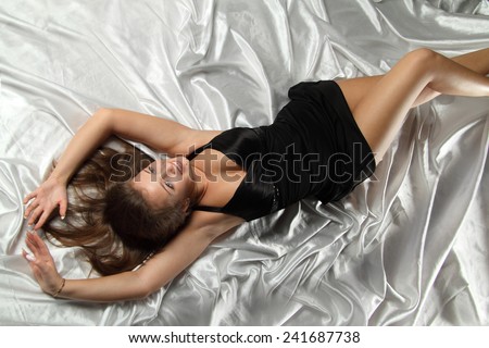 woman in bed on white satin. beautiful makeup, sexy black dress. long hair, long legs, large breasts. satin bedding