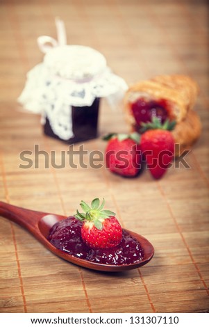 Strawberry jam in a wooden spoon with croissant and a jar of jam