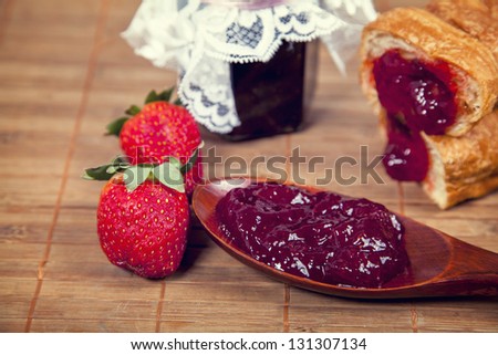 Strawberry jam in a wooden spoon with croissant and a jar of jam