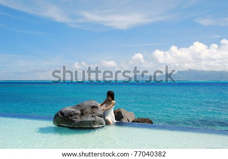 Young lady like a little mermaid , sitting alone between the rocks  in the swimming pool with  scene of Moorea island on the background .