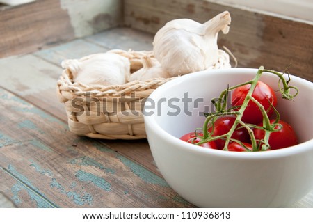 Fresh vine tomatoes in a white bowl along with a basket of fresh garlic. All served on a rustic wooden tray.