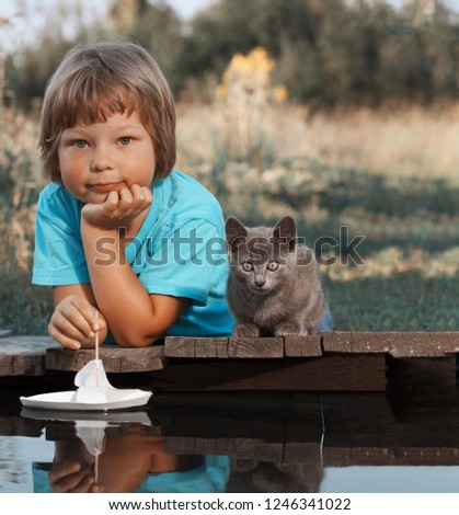 boy and his beloved kitten playing with a boat from pier in a pond summer evening