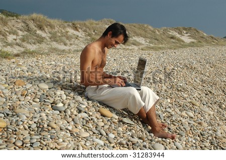 a young man sited on the beach working with a laptop