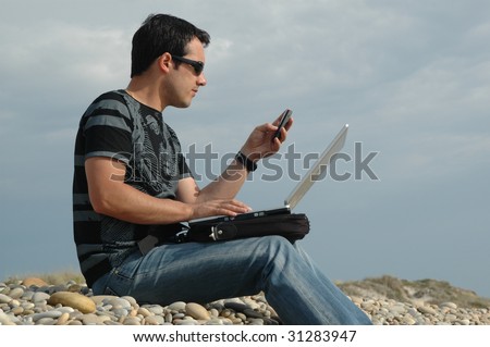a young man sited on the beach working with a laptop and with mobile phone