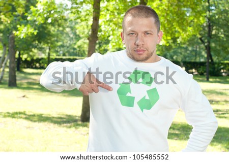 Man standing in a green park and pointing at recycle logo on his shirt