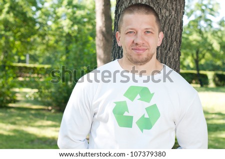 Ecologist with recycling symbol on his shirt standing by the tree, smiling and looking at camera