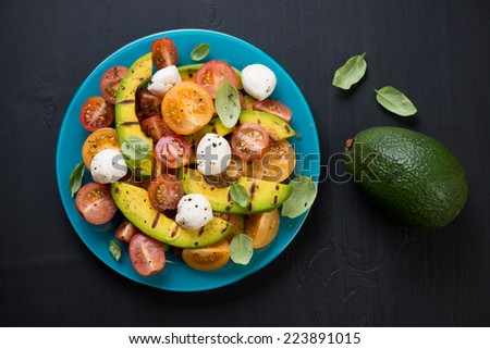 Salad with grilled avocado, tomatoes and mozzarella cheese, black wooden surface, above view