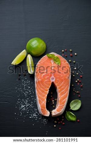 Raw salmon steak with sea salt, pepper, green basil and lime. Black wooden surface, above view