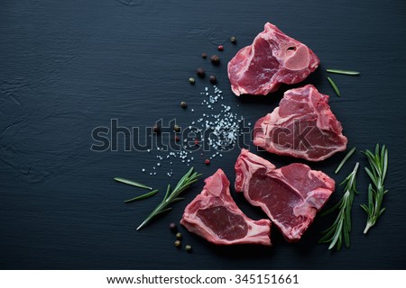 Raw T-bone lamb steaks with seasonings on a black wooden surface