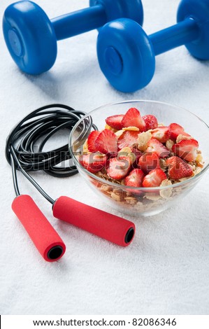 A bowl of muesli with strawberries, a jumping rope and dumbbells, fitness concept