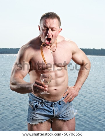 Muscular man with glass of red drink at  lake on sunny day splashing drink