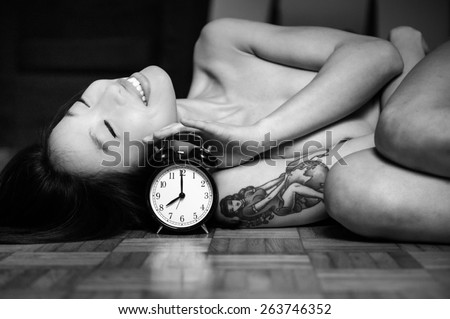 Portrait of nude young asian woman lying and smiling on wooden floor with alarm clock in black and white