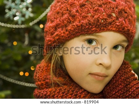 Pretty little girl wearing a red knit hat and matching scarf standing in front of a decorated Christmas Tree.