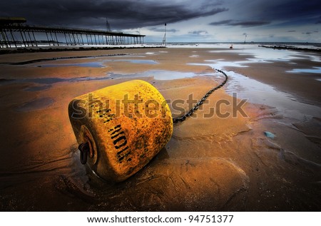 a yellow mooring buoy lying in the sand on the beach waiting for the tide to come in. A moody image with the pier and a threatening sky in the background.