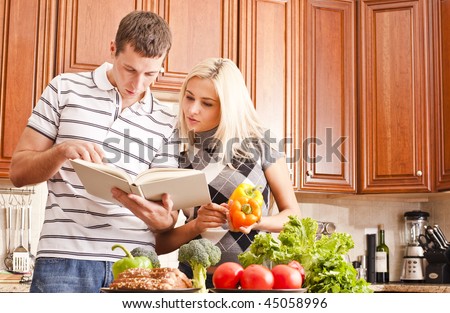 Young couple in the kitchen read an open recipe book. The counter is full of fresh vegetables. Horizontal shot.