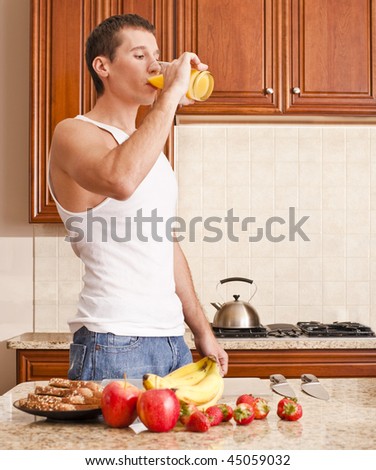Young man wearing tank top and jeans in kitchen drinking a glass of orange juice. Vertical shot.