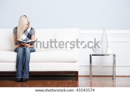 Young woman in checkered shirt and blue jeans sitting on white sofa reading a book. Horizontal shot.