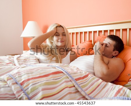 Young couple wake up in bed under striped bedspread. Man is rubbing his eyes while the young woman brushes back her hair. Horizontal shot.