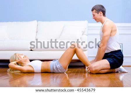 Young woman laughs while doing sit ups on the wood floor.  A young man is holding her feet. Horizontal shot.