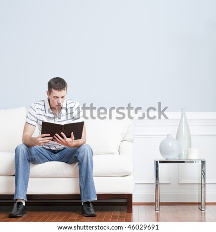 Man sitting and reading on a white couch in his living room. Square format.