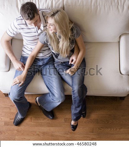 Full length overhead view of affectionate couple sitting together on white love seat. Square format.