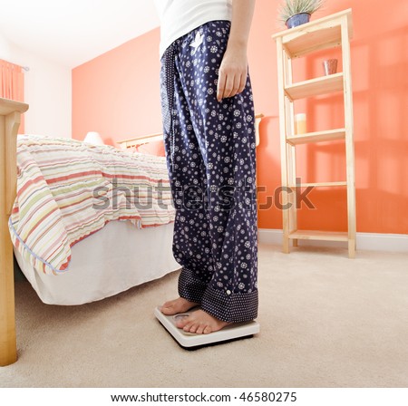 Cropped view of woman standing on a scale next to her bed. Square format.