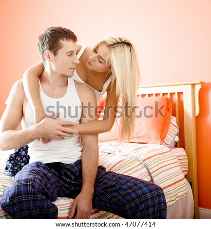Man sits on bed as woman kneels behind him and hugs him. Square format.