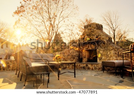 Low-angle view of a flagstone patio with an outdoor stone fireplace and furniture. Rays of sunlight stream down. Horizontal format.