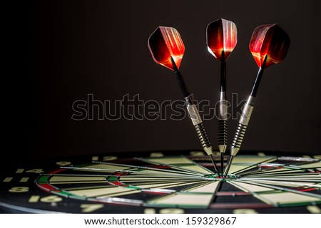Red, black, and green dartboard on its side with three fiery metal tipped darts in the bulls eye. On black background.