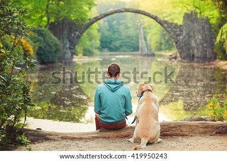 Pensive young man sitting on the pond bank with a his dog - Amazing place in Germany. Rakotzbrucke also known as Devils Bridge in Kromlau.