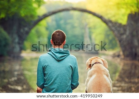 Pensive young man sitting on the pond bank with a his dog - Amazing place in Germany. Rakotzbrucke also known as Devils Bridge in Kromlau.
