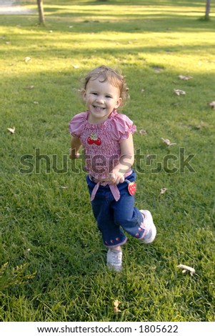 A cute young girl smiles as she runs in the grass at a park.
