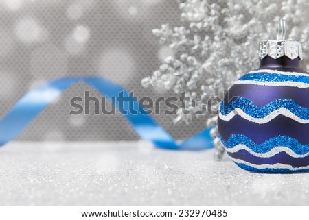 A single blue Christmas ornament sits with a snowflake and ribbon on a glittery surface