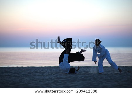 Aikido practice on the beach at sunset; student in white kimono throwing aikido master