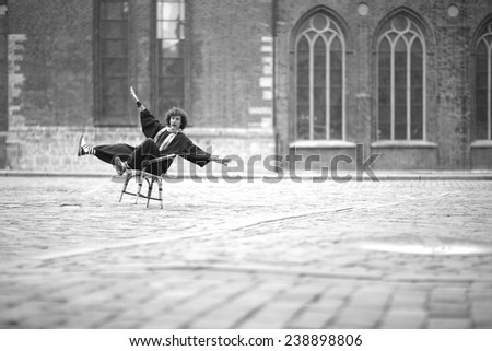 Clown sitting in the middle of the city square, keeping balance by stretching arms and legs, monochrome