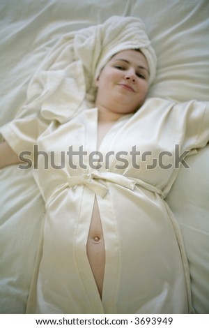 pregnant woman wearing light silk bathrobe and towel around head, lying in bed with bared belly button