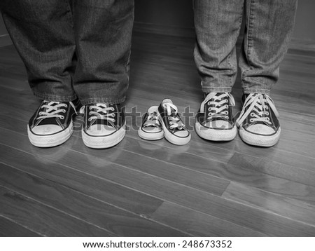 A black and white image of the feet of a man and woman wearing jeans and matching sneakers with a pair of baby sneakers between them