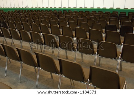 Endless rows of empty brown plastic seats in an exhibition hall, facing a green baize-covered table at the front.