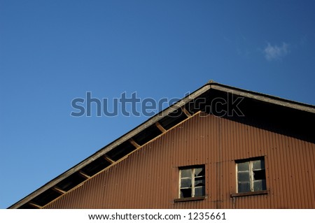 The end of a farmyard barn, two windows on high with broken panes of glass. Space for text in the rich blue sky with a wisp of cloud.