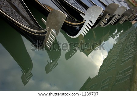 A line of moored gondolas in Venice, their bow decorations reflected in the still waters of the canal.