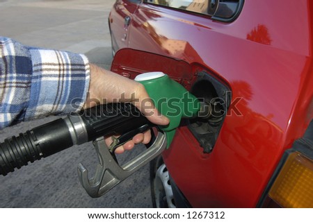 Close up of the hand of a motorist, filling his car with unleaded petrol on a garage forecourt. Beyond is the empty forecourt, but reflected in the car's bodywork is a housing estate and trees.