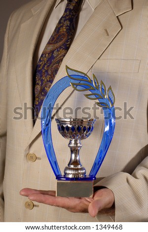 Businessman, dressed in a pale suit, holding an award in the palm of his hand. Space on the plaque of the award for text.