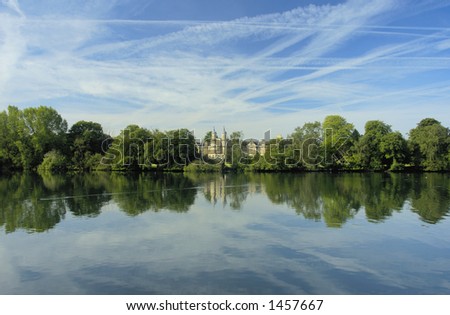 A small castle, hidden amongst the trees, stands on the shore of a calm lake. A waterbird skitters across the smooth surface and the sky, streaked with clouds, is reflected in the water.