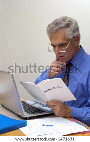 An older businessman gazes in puzzlement at the instructions for his laptop computer, with a hint of worry on his face.