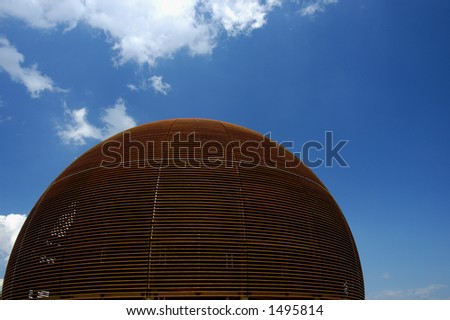 A wooden dome (the Globe of Science and Innovation at the European particle research laboratory CERN, Switzerland) rises against a blue sky with a few fluffy clouds. Space for text on the sky.